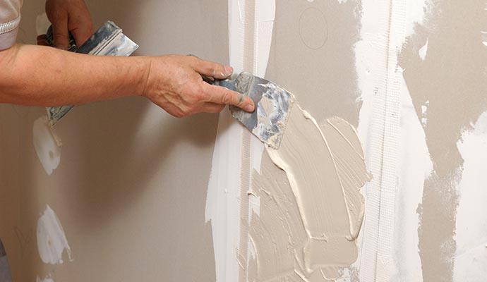 Drywall taping and mudding in progress A close-up of a worker's hands using a putty knife to apply joint compound over mesh tape on a drywall seam.