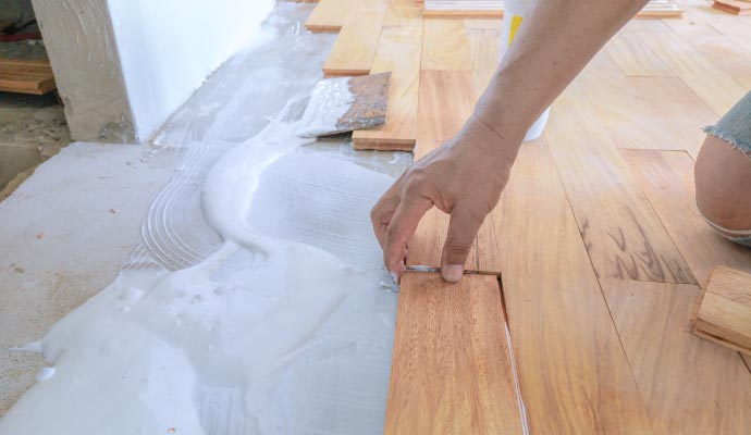 Hardwood floor installation A close-up of a worker's hand laying a hardwood floor plank into a thick layer of white adhesive on a concrete subfloor.
