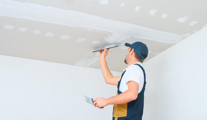 Ceiling drywall finishing A worker in overalls and a cap uses a taping knife to apply joint compound to the seams of a drywall ceiling.